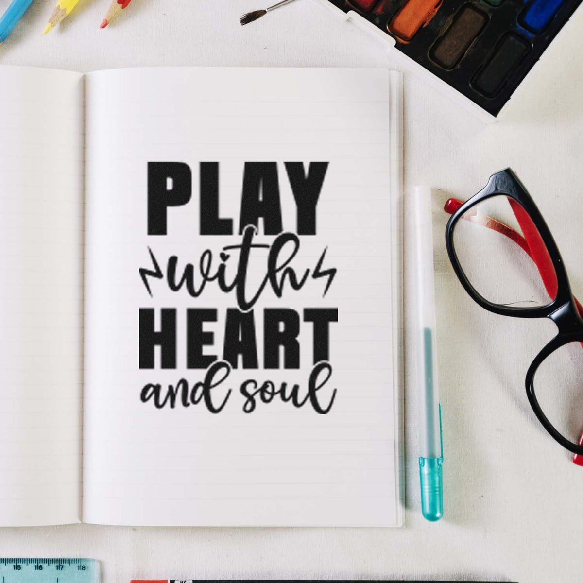 Black rubber stamp reading Play with Heart and Soul in an open notebook, with glasses and a watercolor palette.