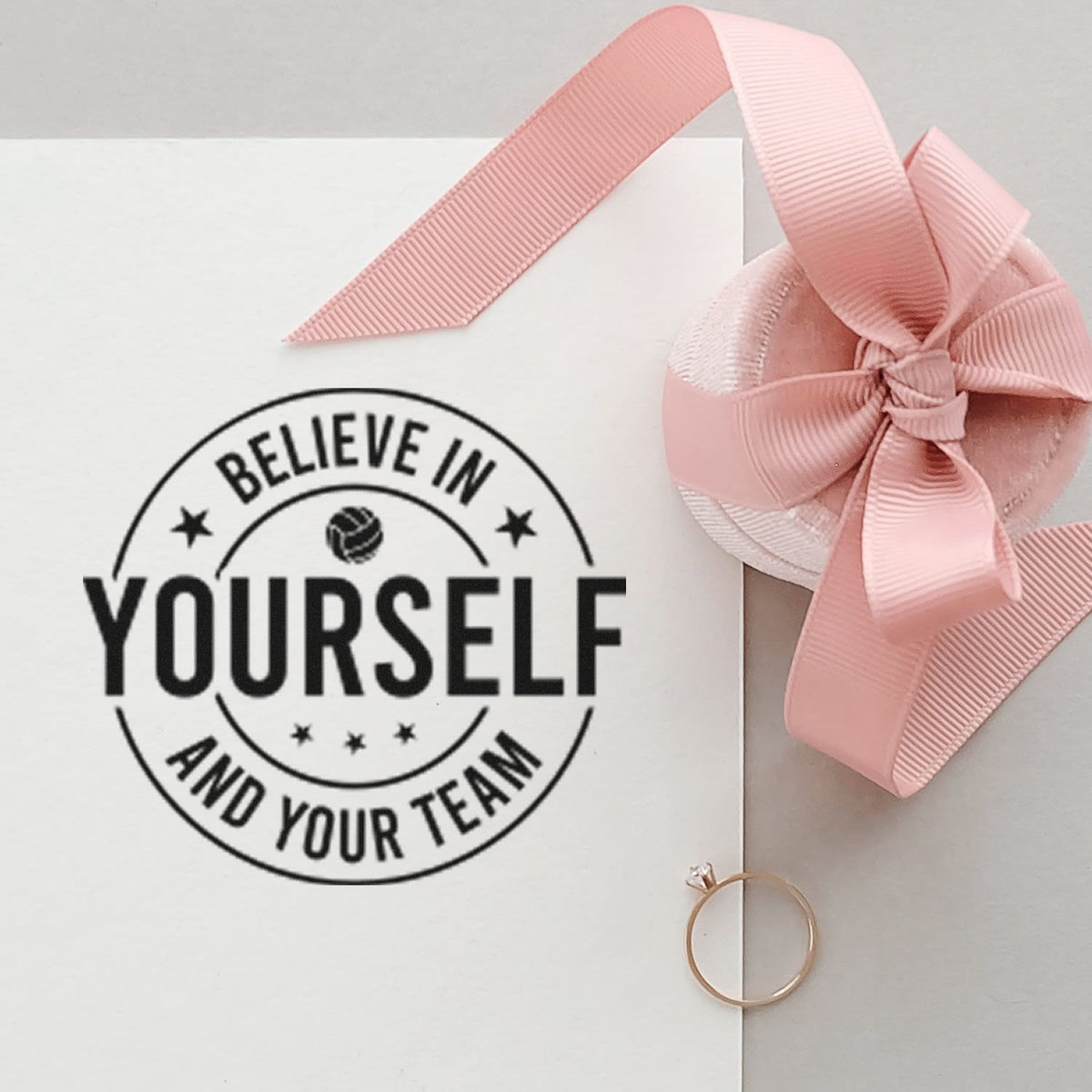 White stamp and seal card with black Believe in Yourself and Your Team seal, pink ribbon bow, and rose-gold ring.