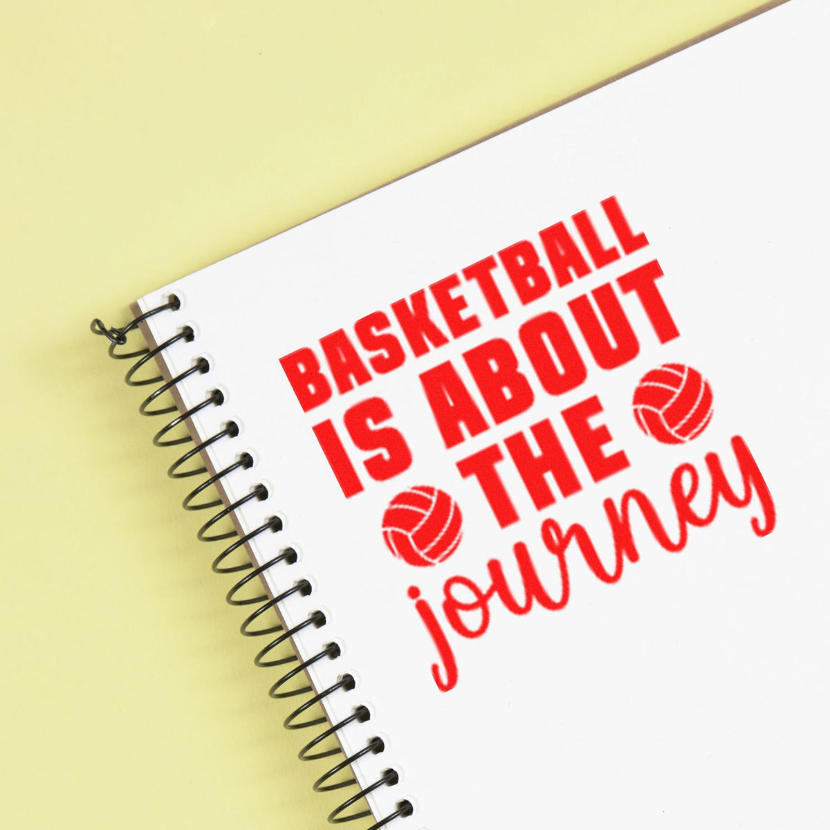 Red basketball stamp and seal on white spiral notebook over pale yellow background, reading Basketball is about the journey.