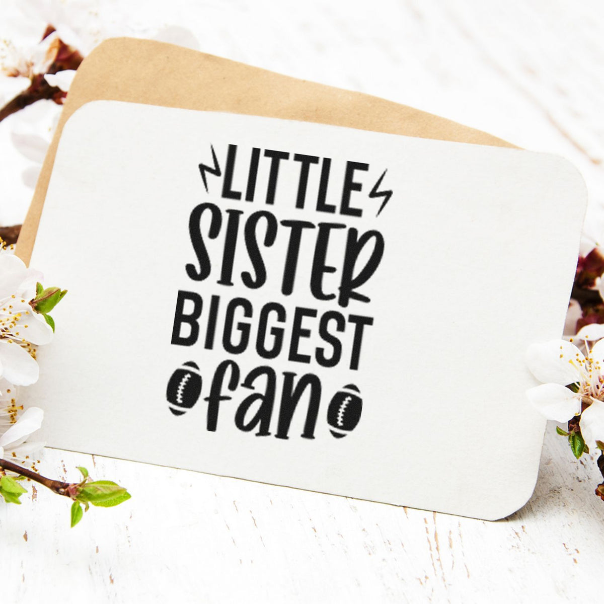 White stamp and seal card on tan backing with black Little Sister Biggest Fan text and football icons, framed by blossoms.