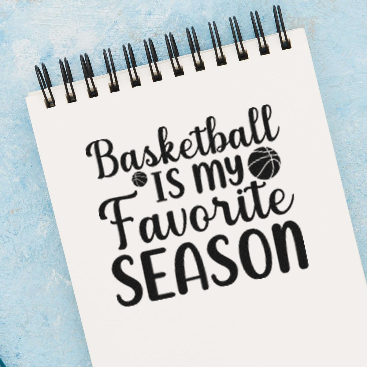 Black basketball stamp and seal design on white spiral notepad, reading Basketball Is My Favorite Season, on blue background.