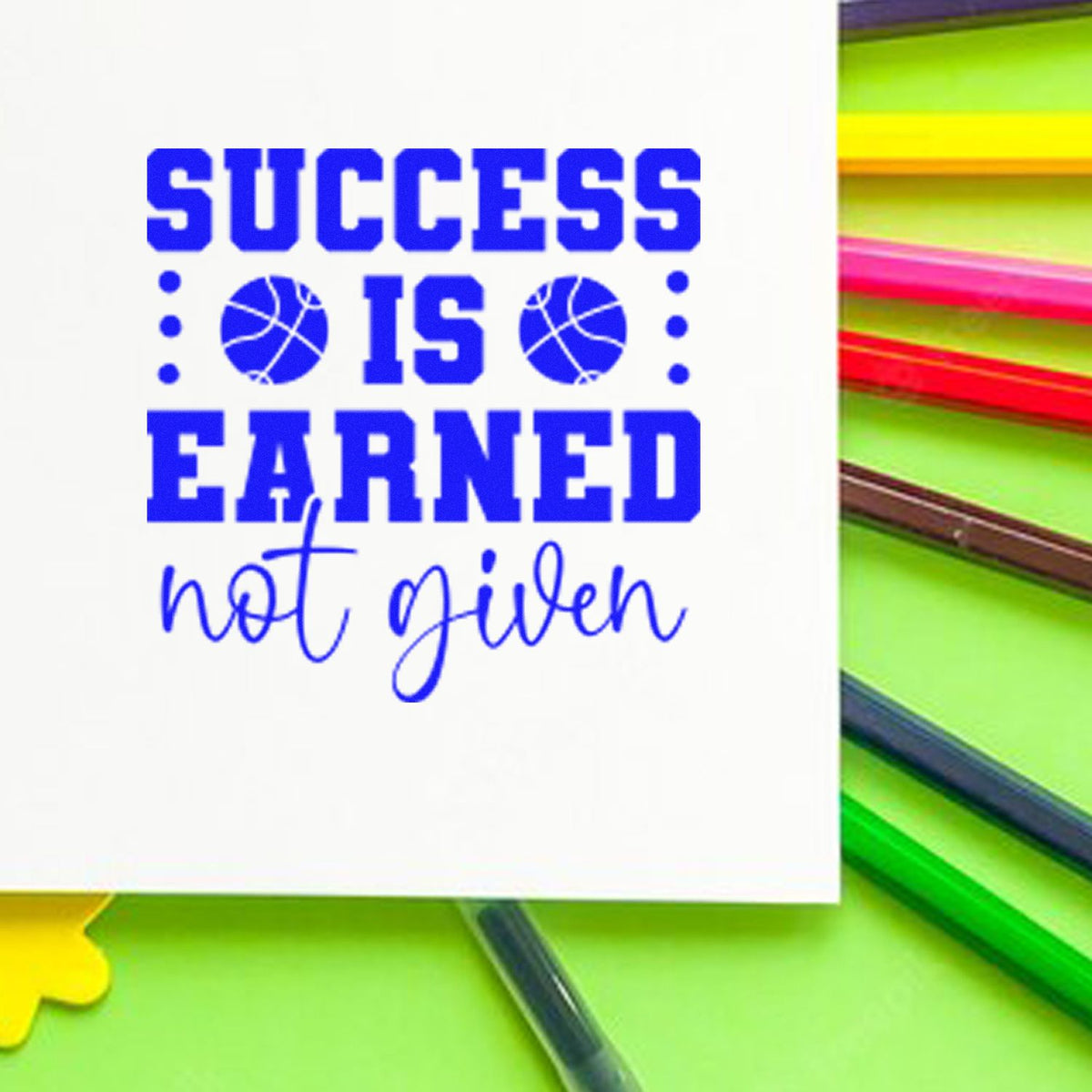 Blue motivational stamp/seal reading Success Is Earned, Not Given on white card, with neon green, pink, and yellow pens.