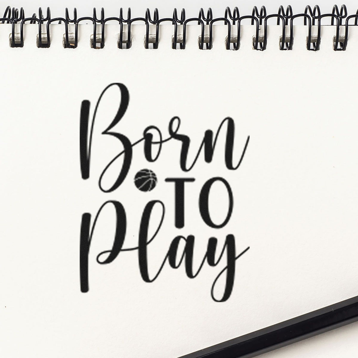 Black stamp and seal design on white spiral notebook, reading Born to Play with a basketball icon.