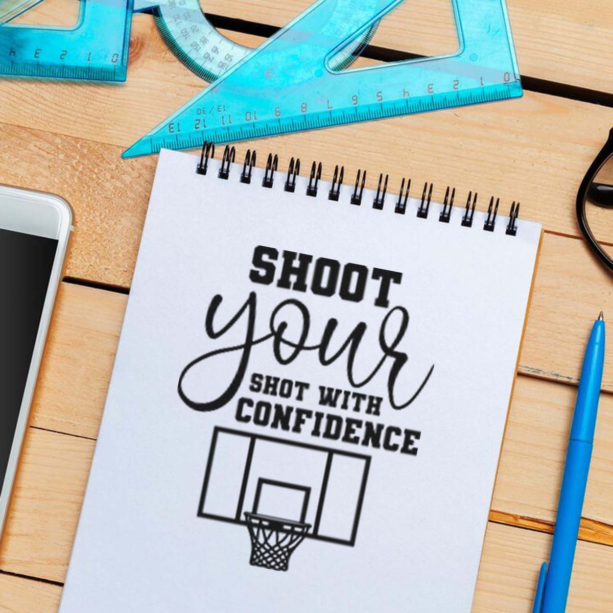 Custom stamp and seal product design on white spiral notebook, with black basketball graphic, turquoise rulers, pen, and wood desk.