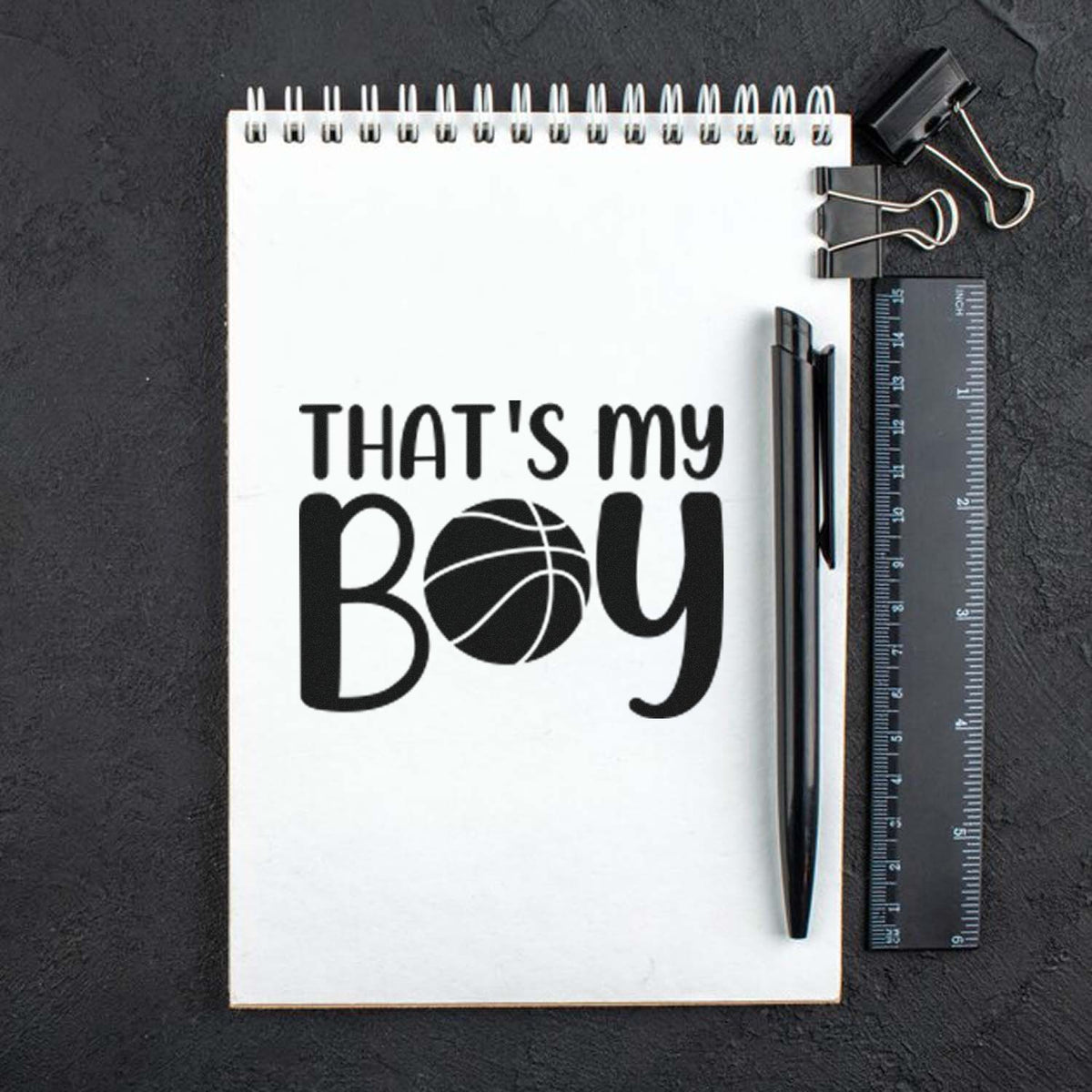 White spiral notebook with black THATS my BOY basketball stamp, beside black pen, ruler, and binder clips on dark background.