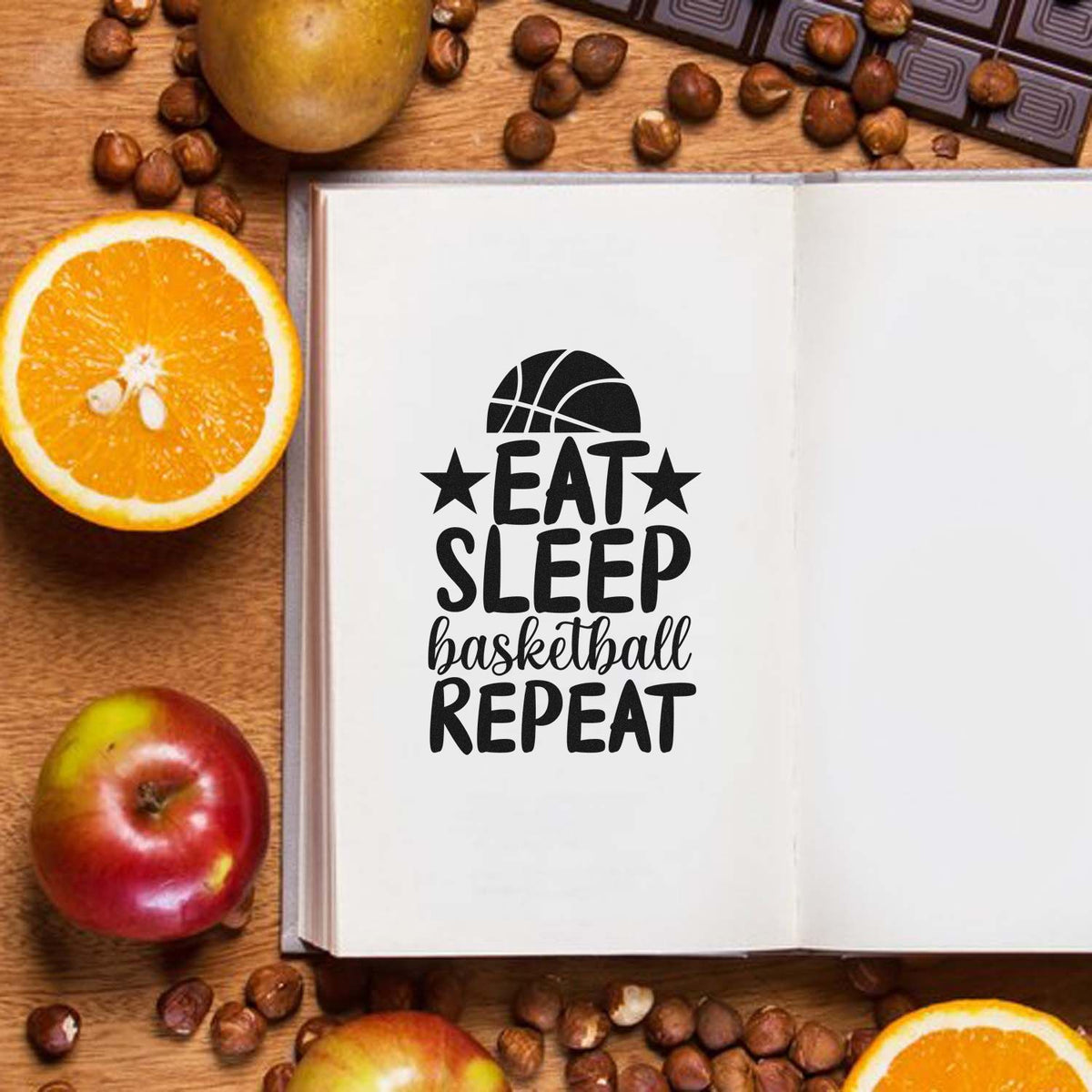 Black basketball stamp seal reading Eat Sleep Basketball Repeat on an open white notebook, with orange slices, apples, and coffee beans.
