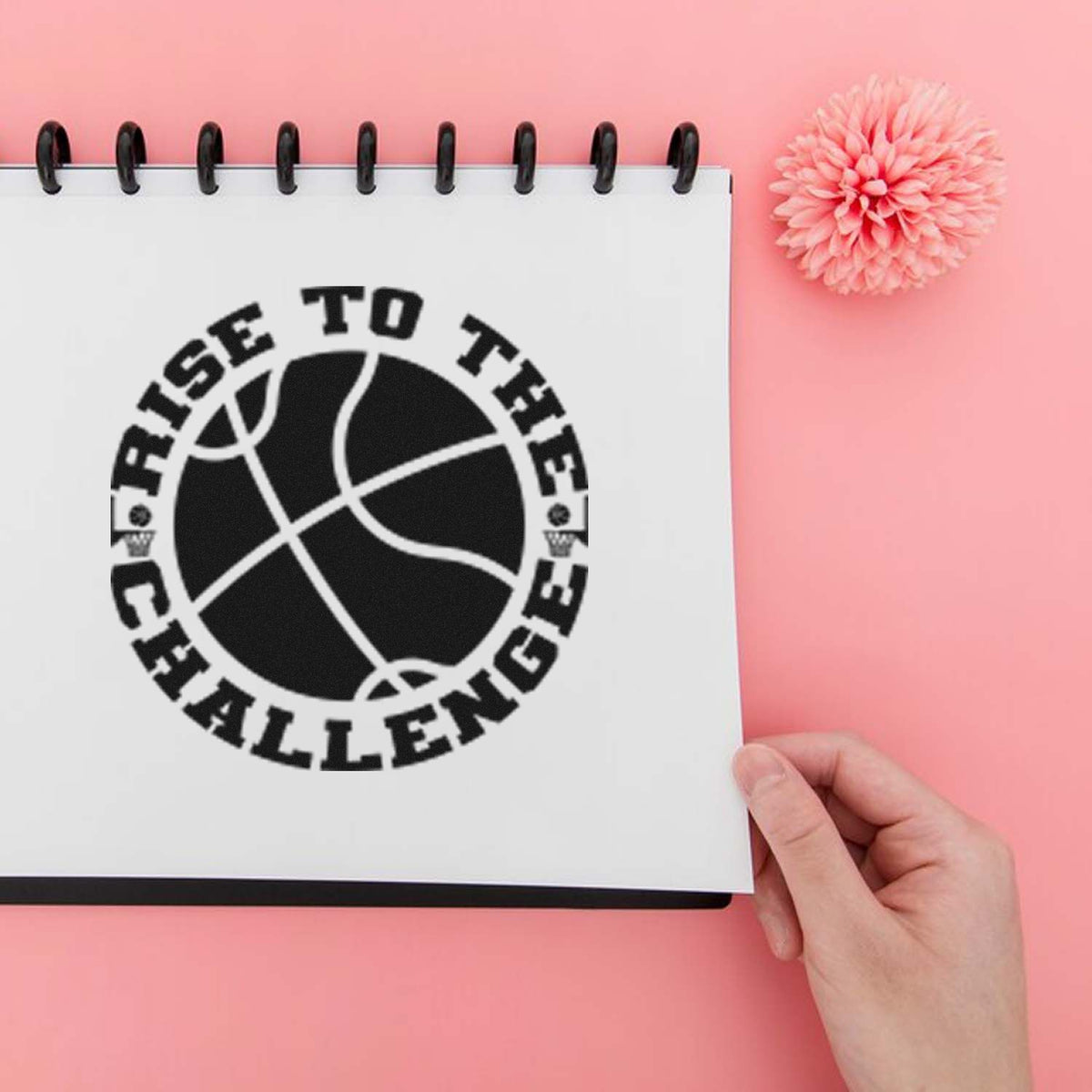 Black basketball Rise to the Challenge stamp and seal on a white spiral notebook, with a pink background and pink flower.