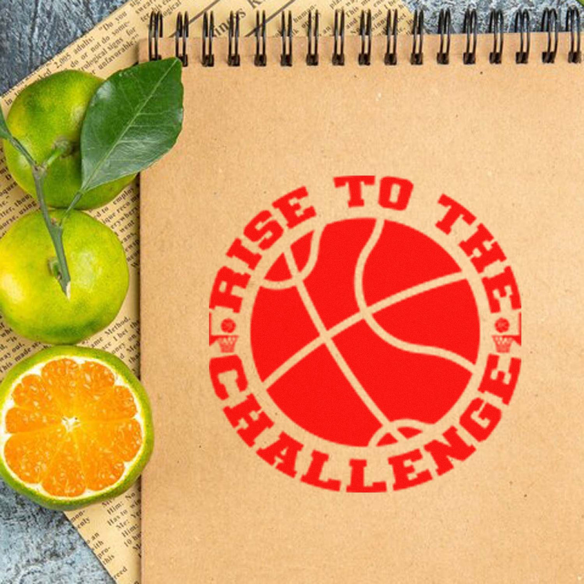 Kraft spiral notebook with red RISE TO THE CHALLENGE basketball stamp seal, beside green oranges and a sliced orange.