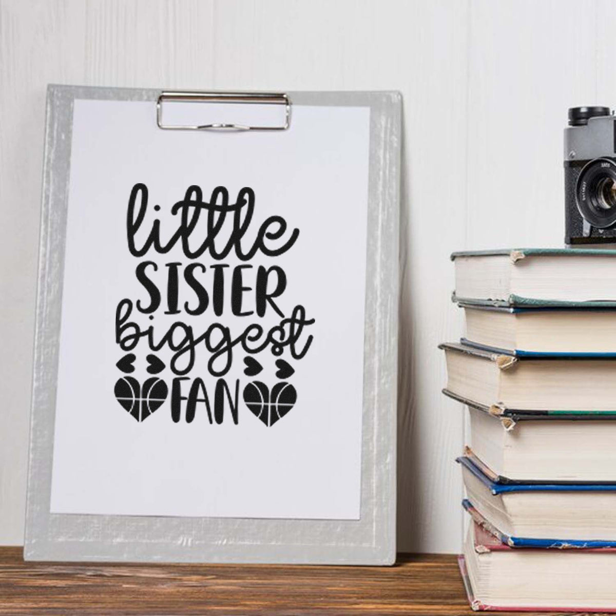 Black little sister biggest fan stamp/seal design on a white clipboard beside stacked books and a vintage camera.
