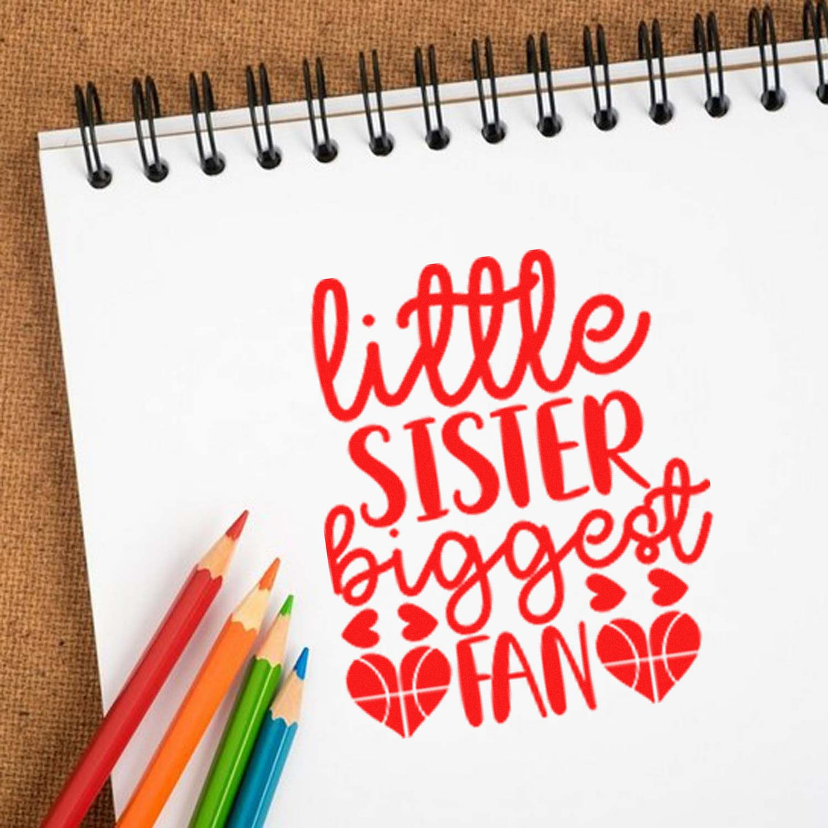 Red little sister biggest fan stamp and heart seal design on white spiral notebook, with colored pencils.