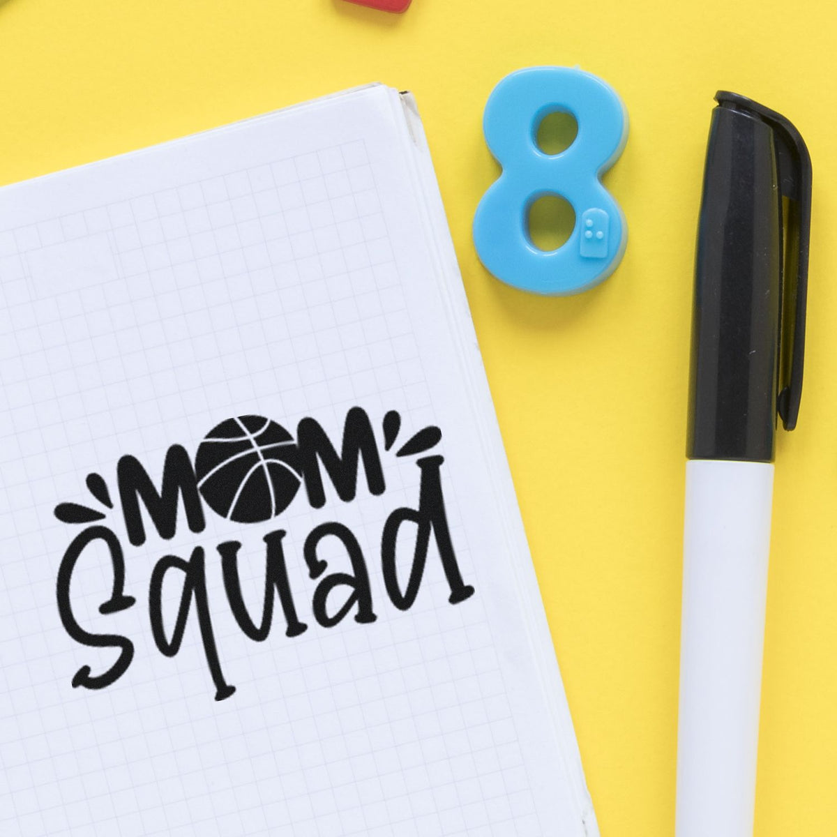 Black Mom Squad stamp on graph notebook, blue 8 seal stamp, and black marker on bright yellow background.