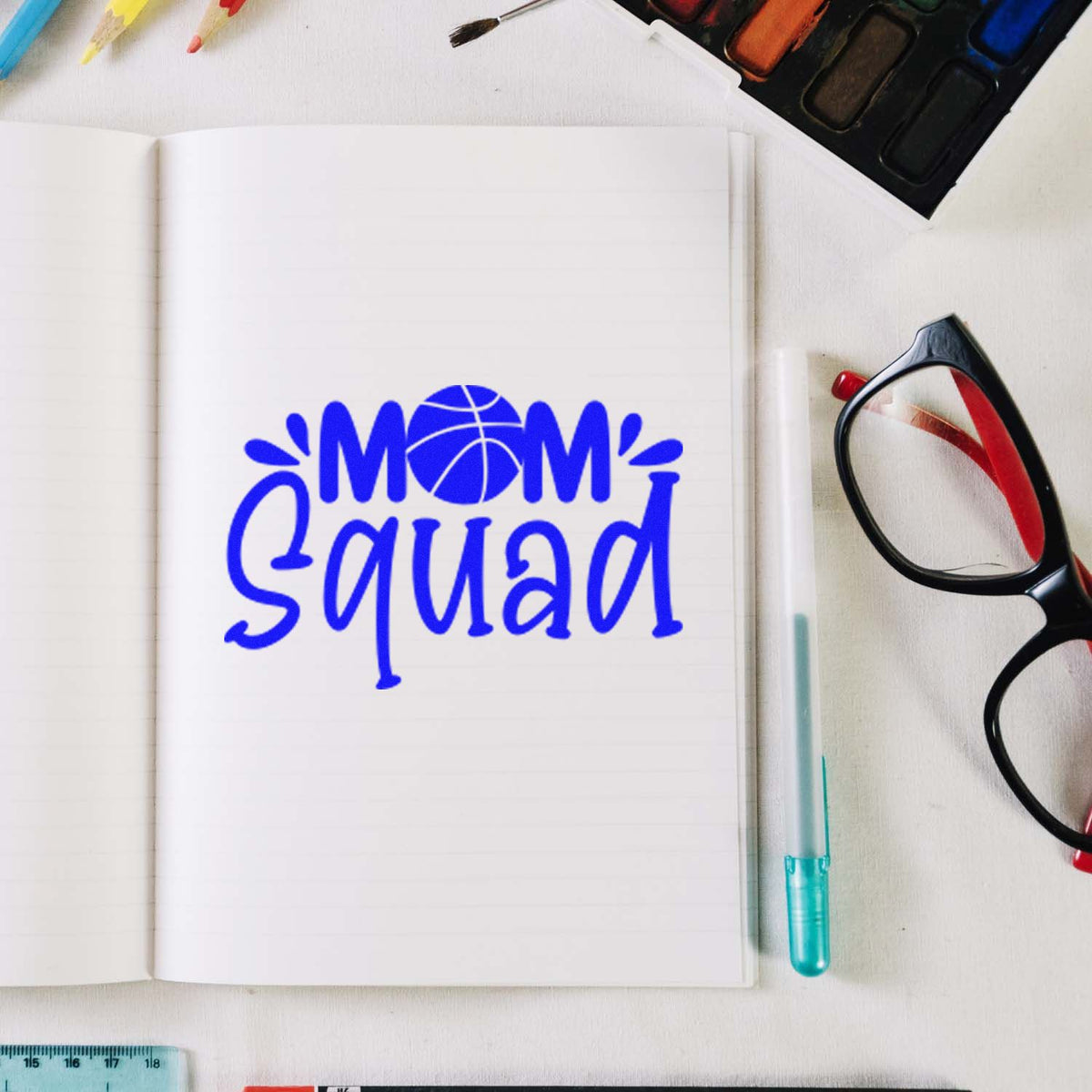 Blue Mom Squad stamp on open notebook, with seal products, glasses, and art supplies on a white desk.