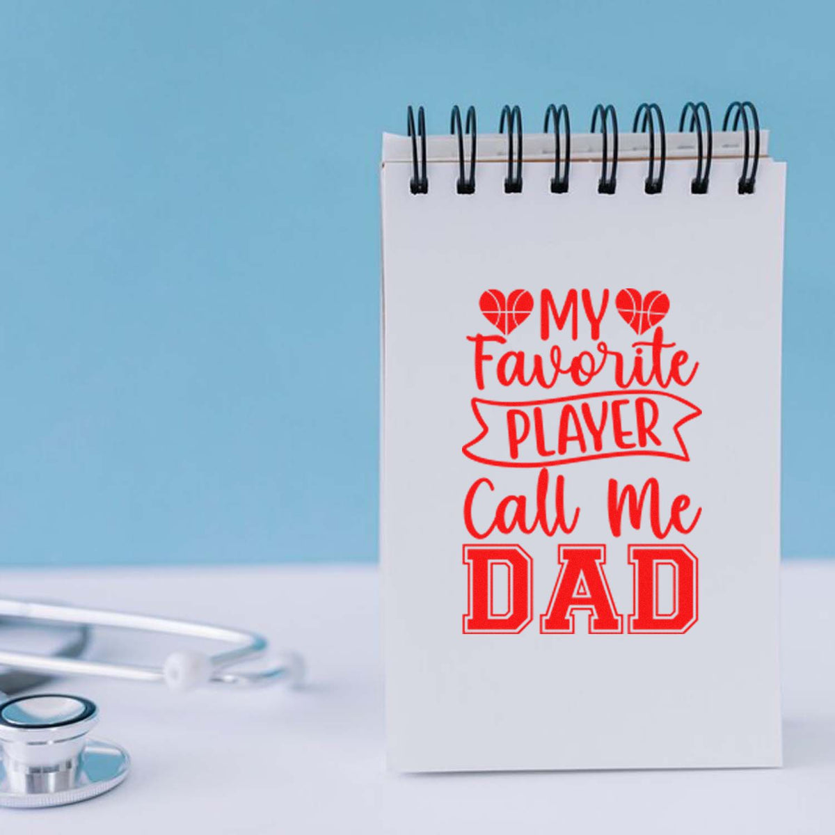 Custom red stamp design on white spiral notebook reading My Favorite Player Call Me Dad, with hearts and a stethoscope.
