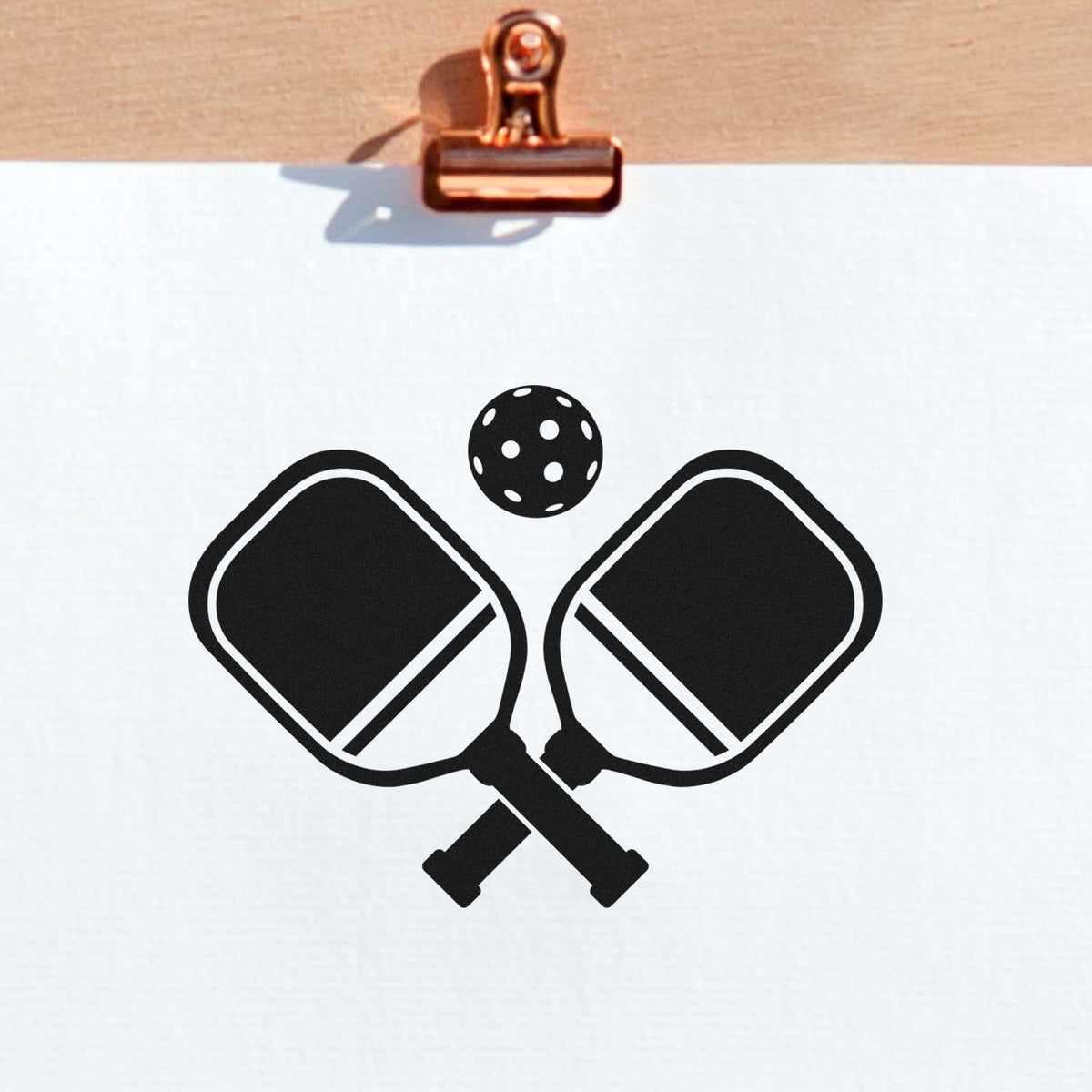 Black pickleball paddle and ball stamp design on white paper, clipped by a copper clipboard clip.