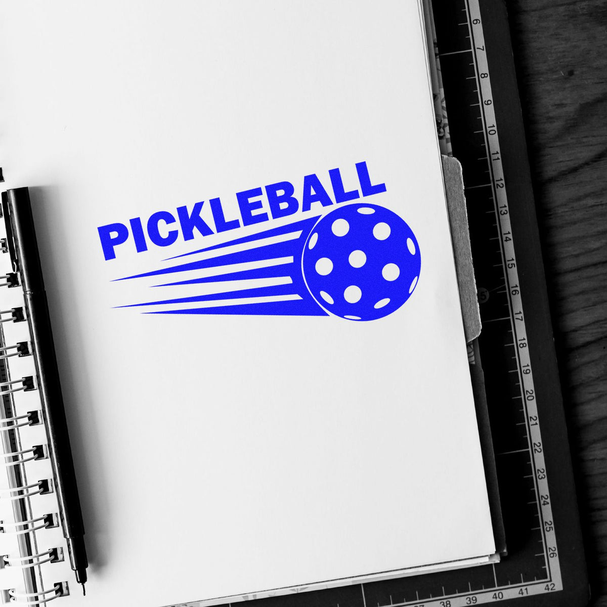 Blue pickleball stamp/seal design with ball graphic and speed lines on white page, beside a black pen and cutting mat.