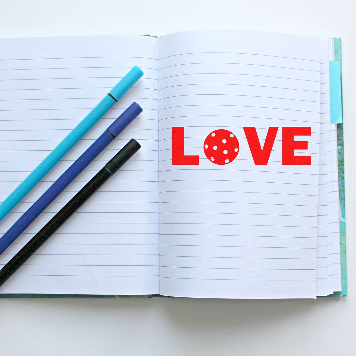 Three stamp and seal penscyan, purple, and blackbeside a red LOVE design on an open lined notebook.