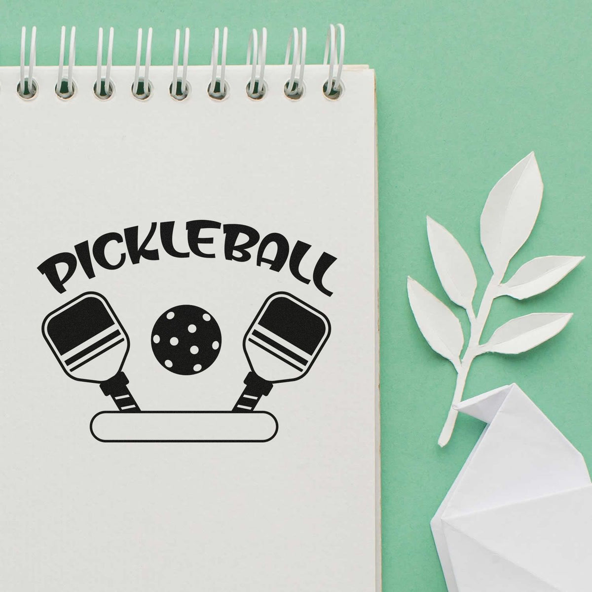 Pickleball stamp and seal set with black paddles and ball on a white spiral notebook against a mint green background.