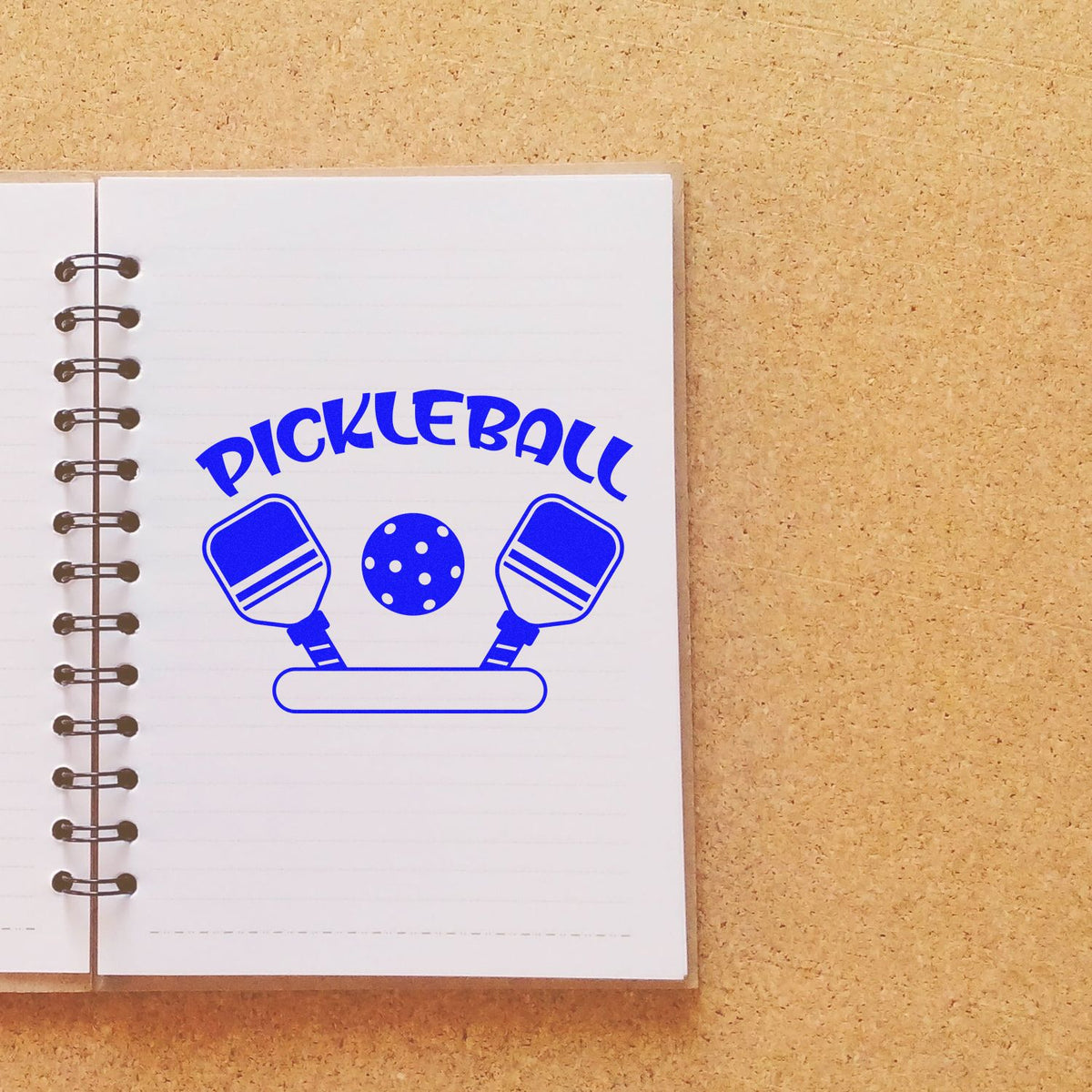 Blue pickleball stamp and seal design on a white spiral notebook, with two paddles, ball, and bold arched text.