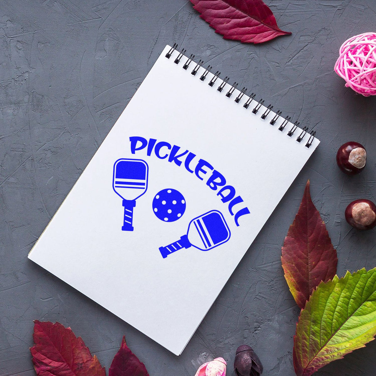 Blue pickleball stamp and seal on white spiral notebook, with paddle icons and ball on gray surface with autumn leaves.