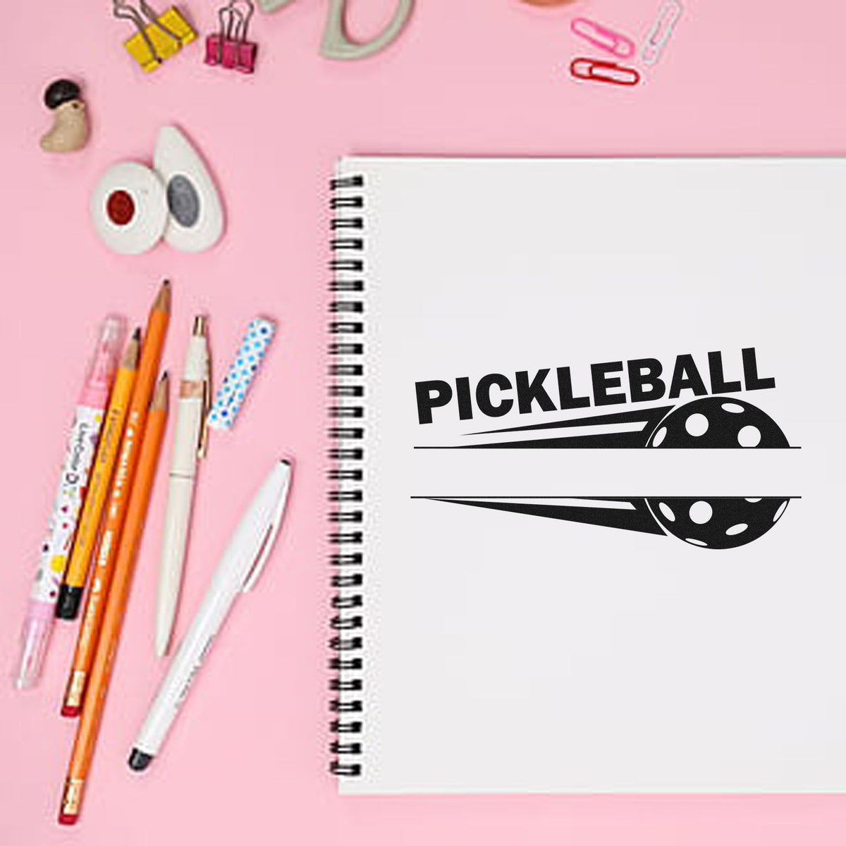 Black pickleball stamp and seal on white spiral notebook, surrounded by pens, clips, and pastel accessories on pink background.
