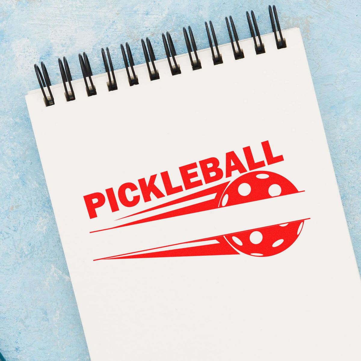 Red pickleball stamp and seal on white spiral notepad with black coil binding and blue background.
