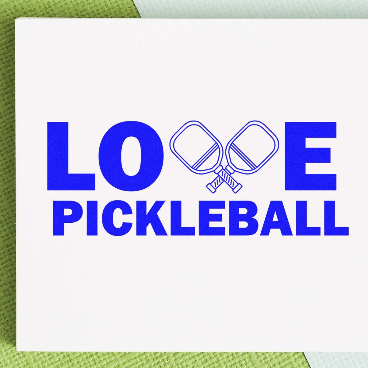 Blue LOVE PICKLEBALL stamp with crossed paddle seal on a white card over a green textured background.