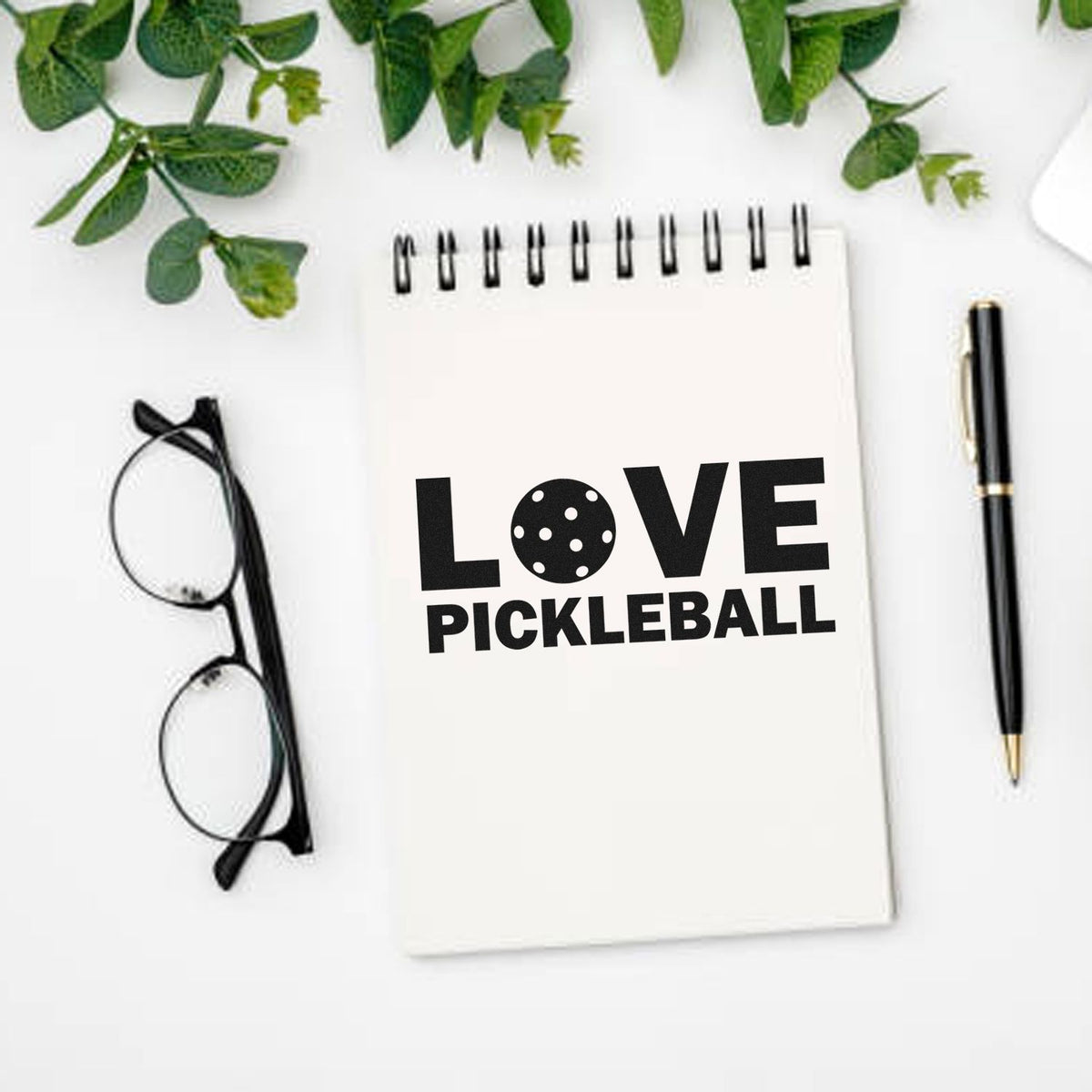 Custom stamp/seal design: black LOVE PICKLEBALL on white spiral notebook, with glasses, pen and green leaves.