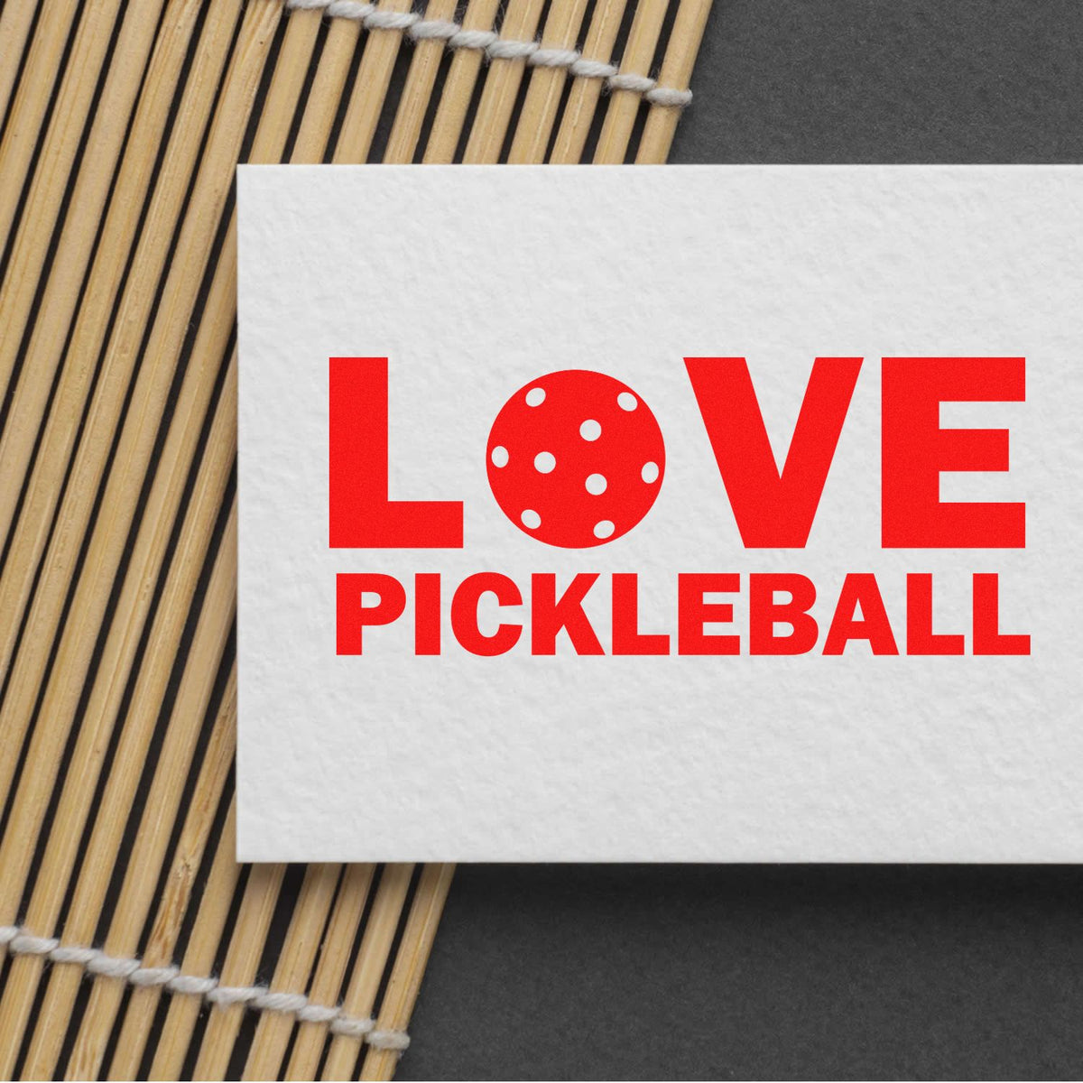 Red LOVE PICKLEBALL stamp on white card, with dot-pattern pickleball, on bamboo mat over dark gray background.