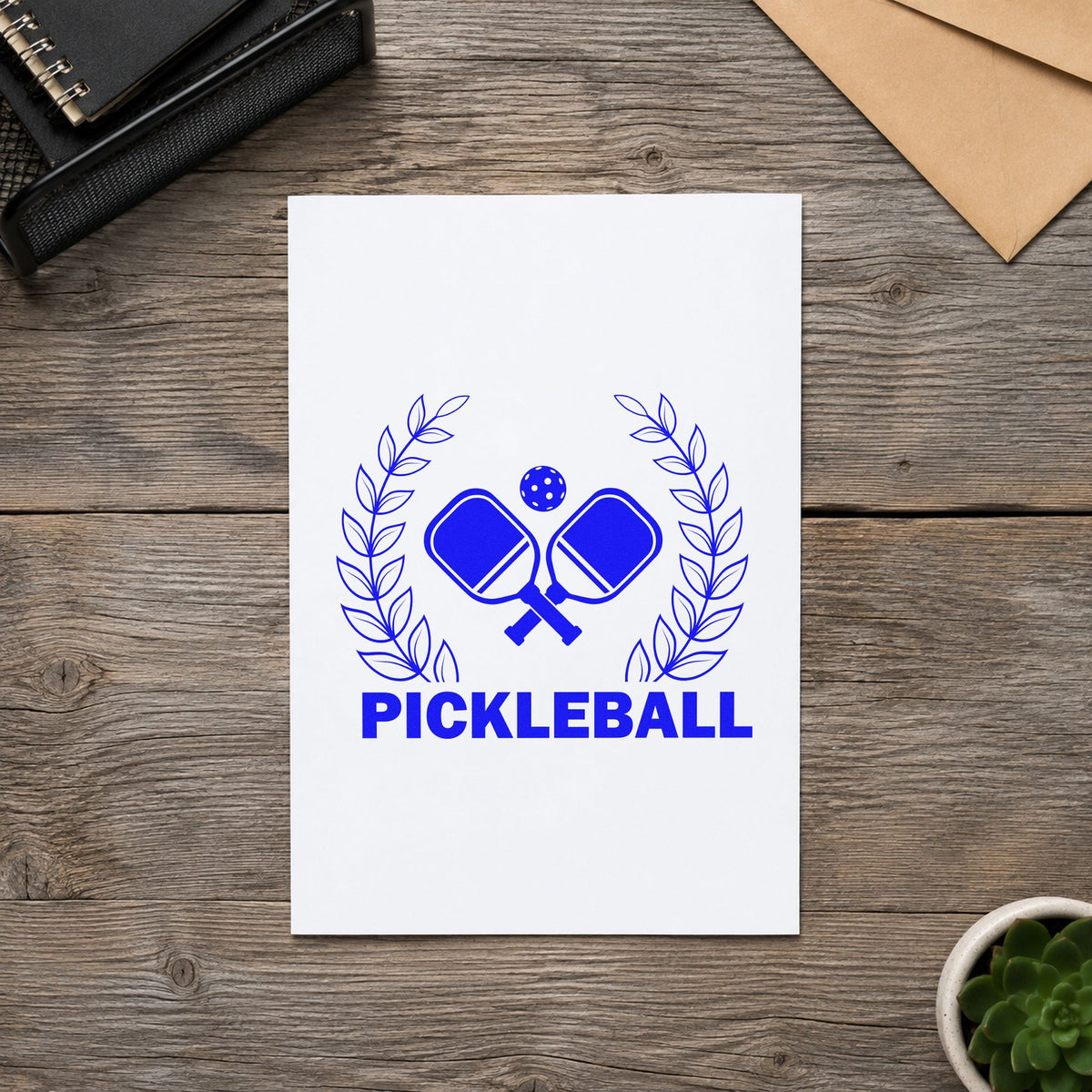 Pickleball Laurel Paddle Badge craft stamp in blue ink on cardstock, an easy option for warmup jacket printing.