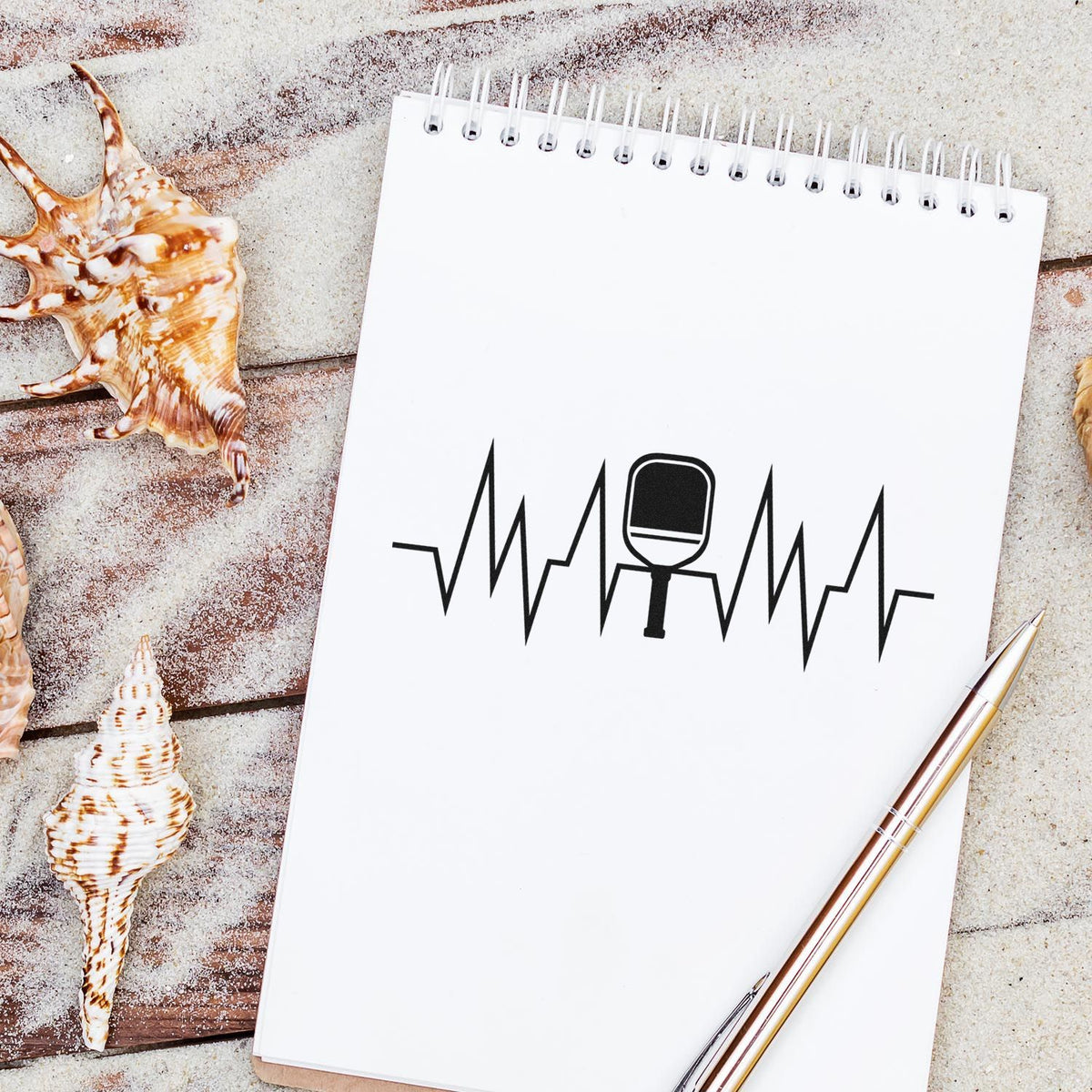 White spiral notepad featuring a black stamp-and-seal logo, with seashells, sandy backdrop, and a gold pen.