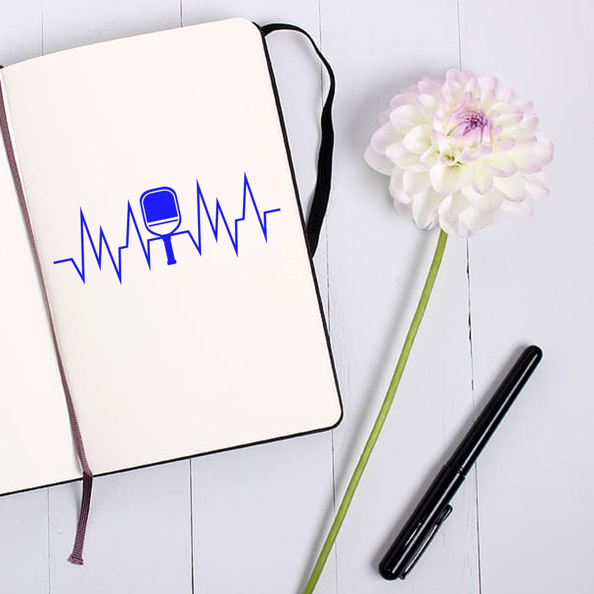 White notebook with blue stamp-and-seal logo, black pen, and white flower on a pale wood background.
