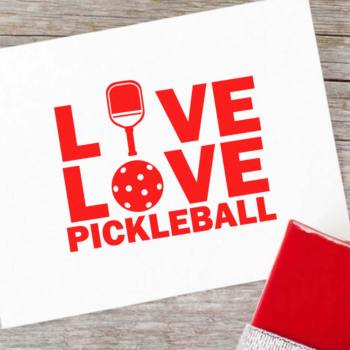 Red LOVE PICKLEBALL stamp design with paddle and ball icons on white card, beside a red seal block on wood background