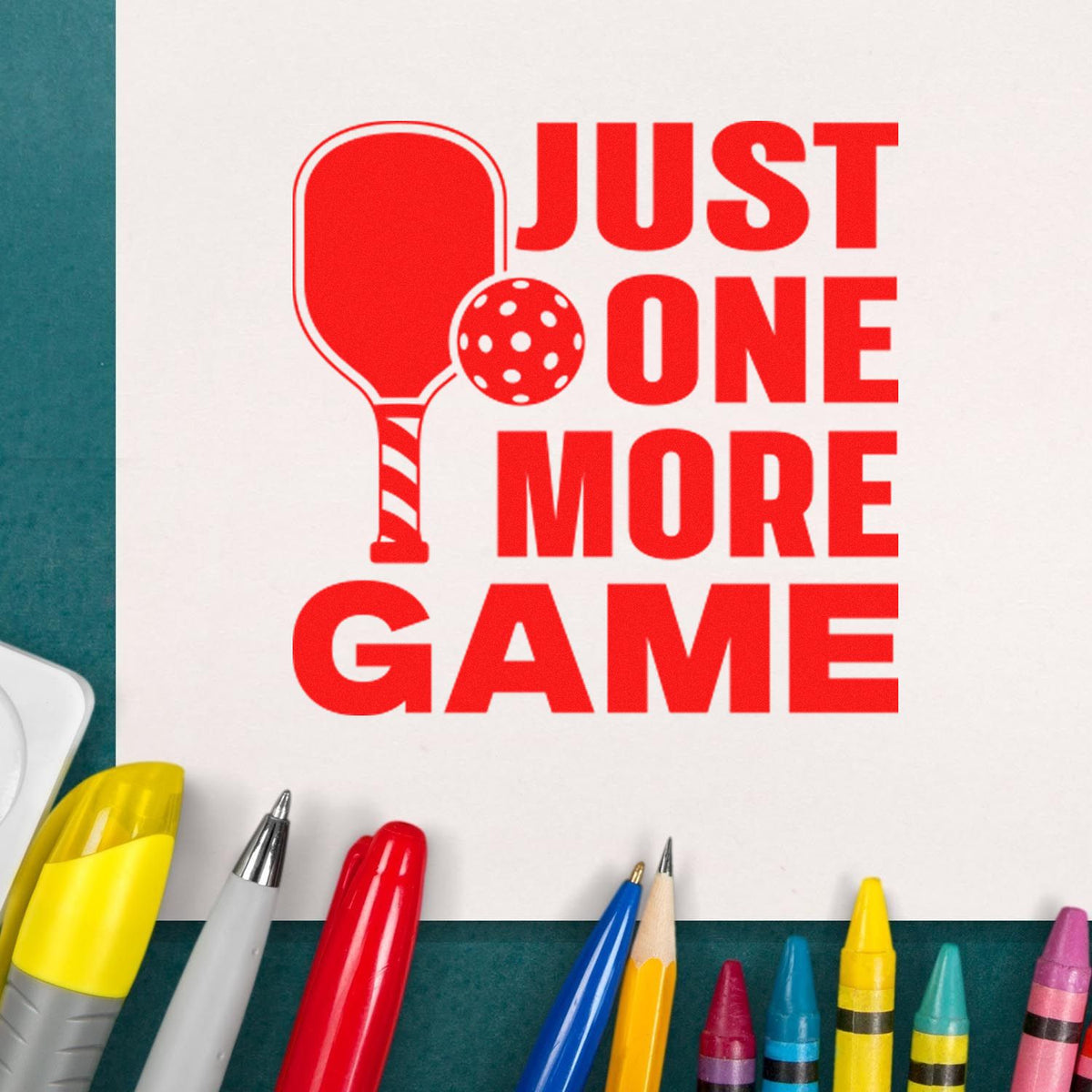 Red stamp-style pickleball seal reading JUST ONE MORE GAME on white paper, with colorful pens.