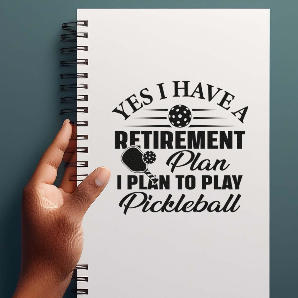 White spiral notebook with black stamp-seal retirement plan text, pickleball paddle graphic, on a teal background.