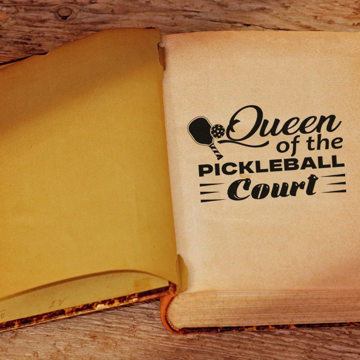 Black pickleball stamp seal reading Queen of the Pickleball Court on cream paper with yellow page spread.