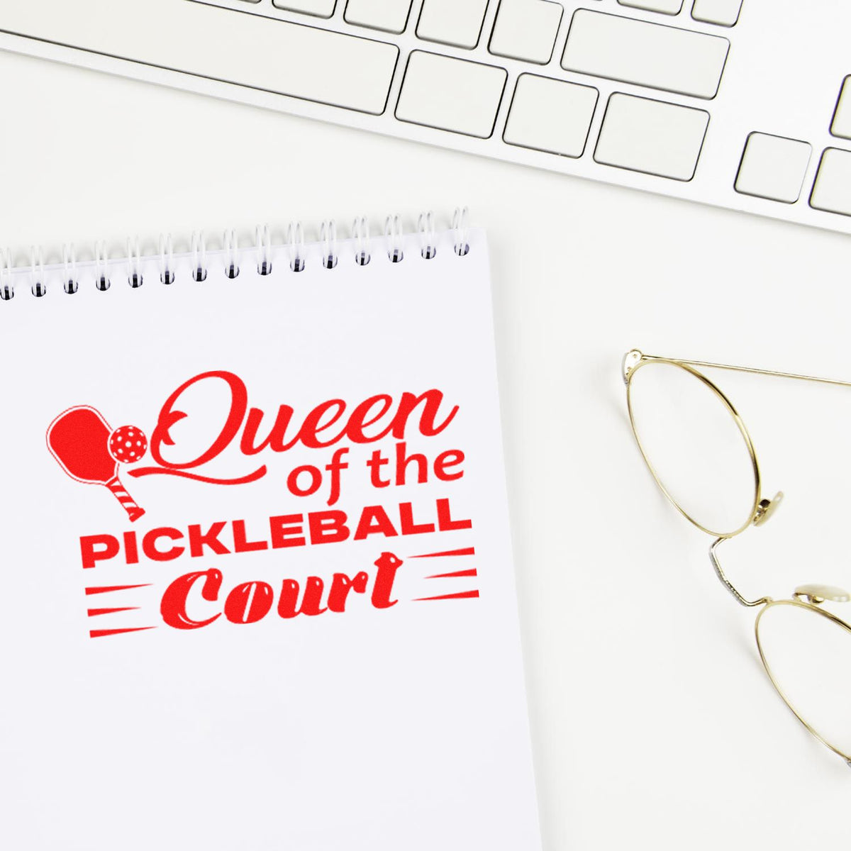 Custom stamp and seal on white notepad, red Queen of the Pickleball Court design beside gold eyeglasses and keyboard.
