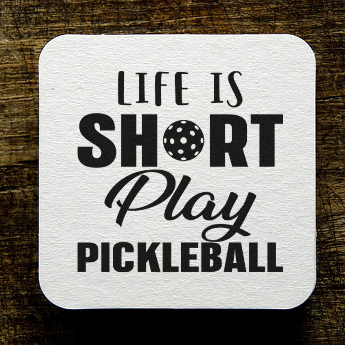 White square pickleball stamp seal with black text, Life Is Short Play Pickleball, on a brown wood background.