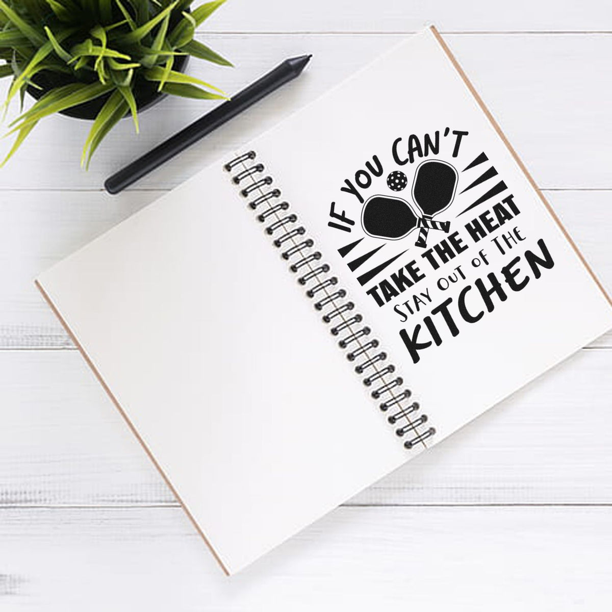 Black kitchen quote stamp-and-seal on a white spiral notebook, with pen and green plant, in bold black lettering.