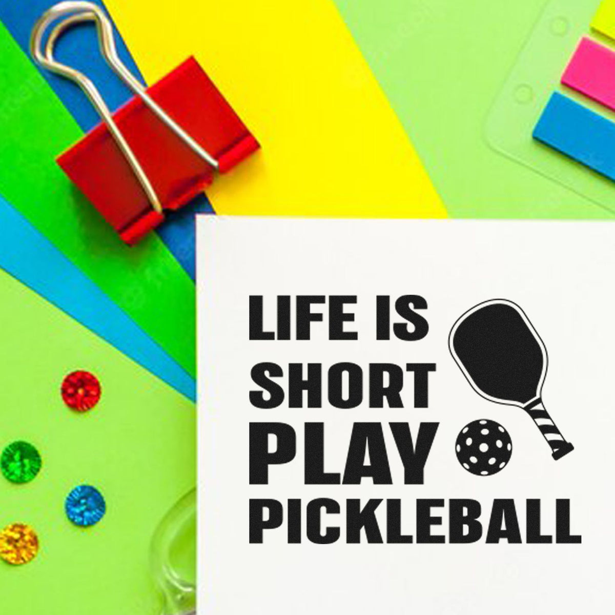 Colorful stamp and seal products with a red binder clip, neon paper, and a white pickleball stamp reading Life is short play pickleball.