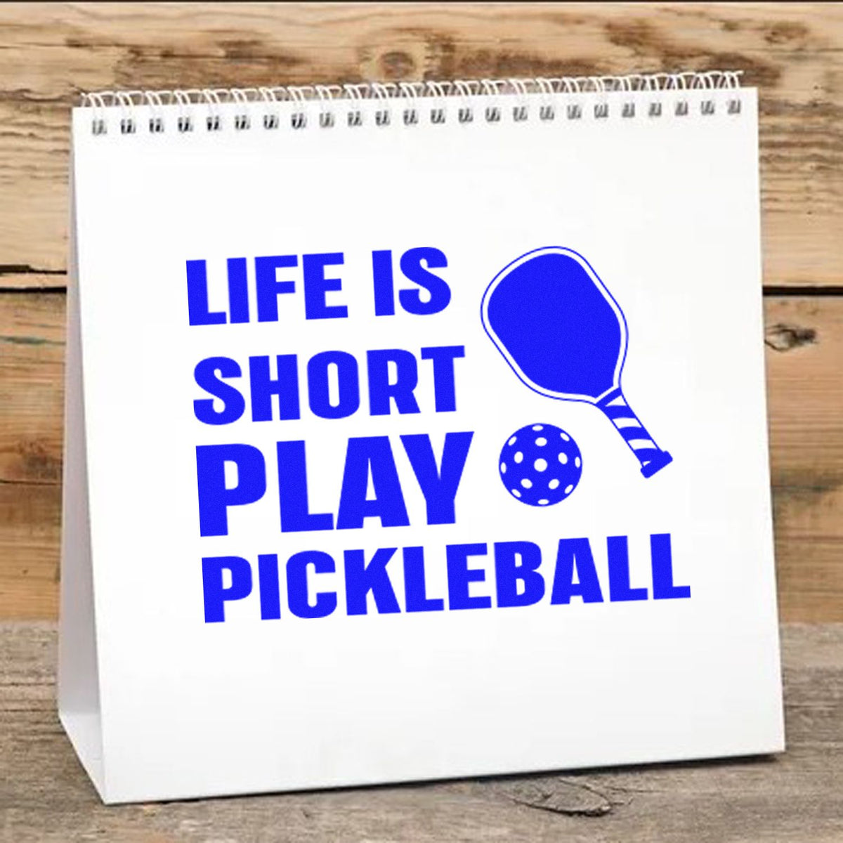 Blue pickleball stamp and seal design on a white spiral-bound bag, with paddle and dotted ball on a wood background.