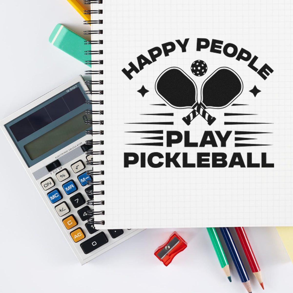 Black pickleball stamp seal on white grid notebook with mint calculator, colorful pencils and sharpener.