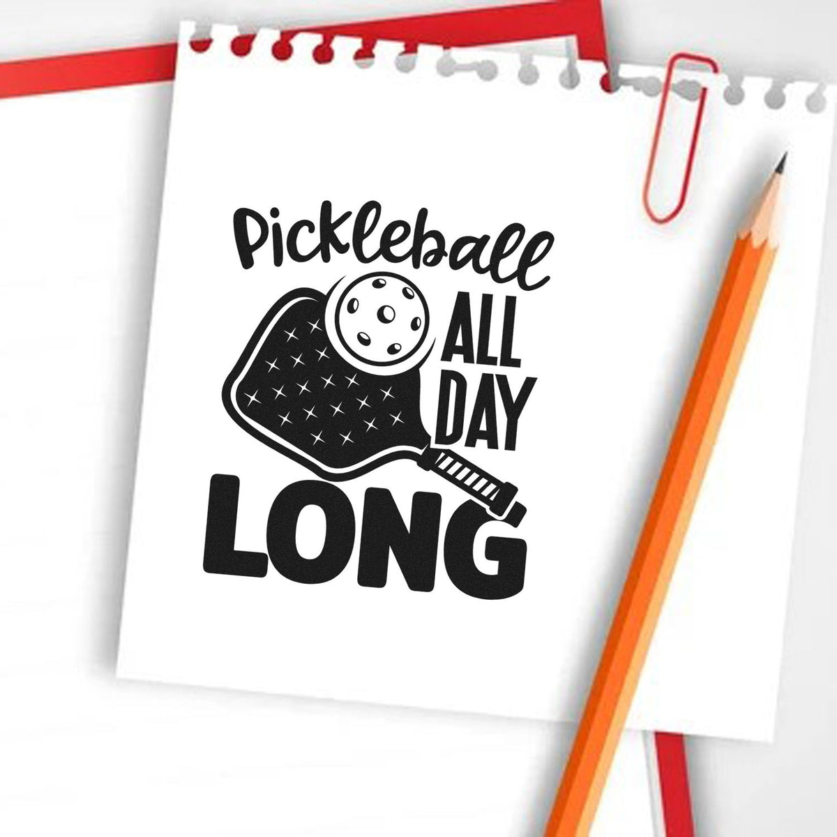 Black pickleball paddle stamp design on white notepad with red paperclip and orange pencil, reading Pickleball All Day Long.