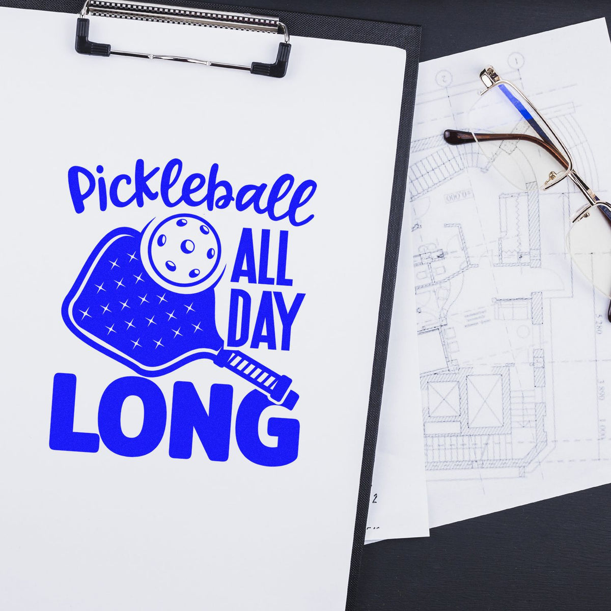 Blue pickleball paddle stamp and seal design on white clipboard, beside eyeglasses and drafting plans.