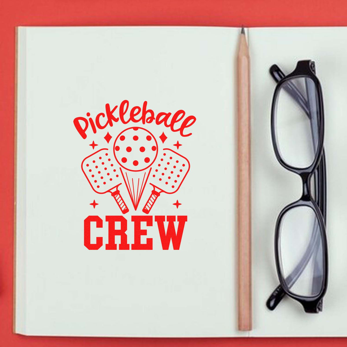 Red pickleball CREW stamp design in a notebook, with paddle and ball seal graphic, beside a pencil and black glasses.