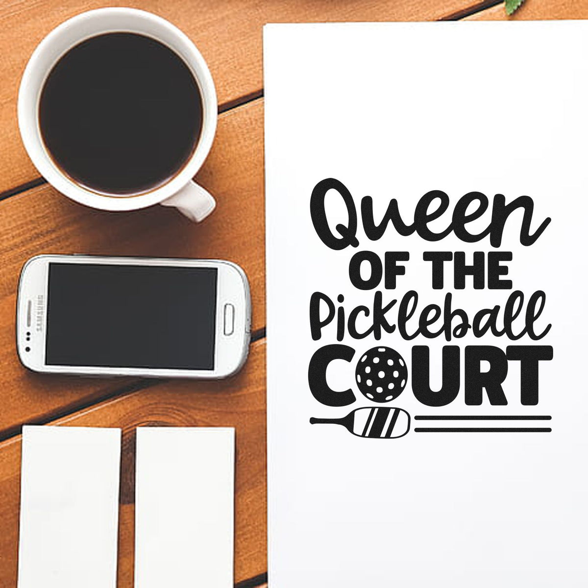 Black pickleball stamp and seal design on white paper reading Queen of the Pickleball Court beside a coffee cup.