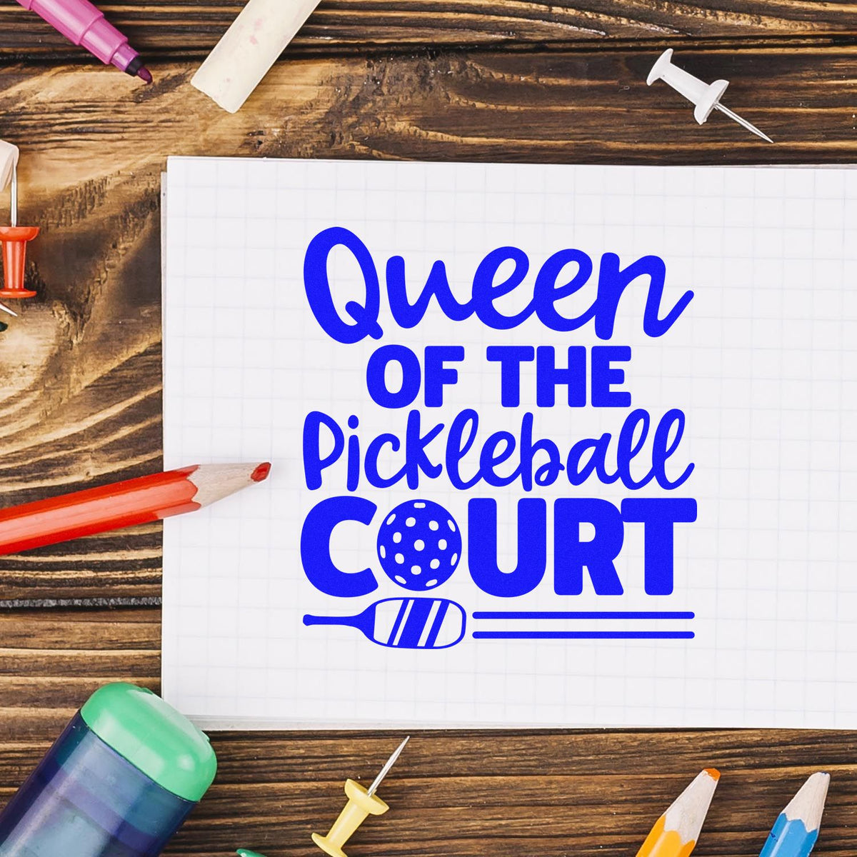Blue Queen of the Pickleball Court stamp design on white grid paper, with colorful pens and push pins on a wood desk.