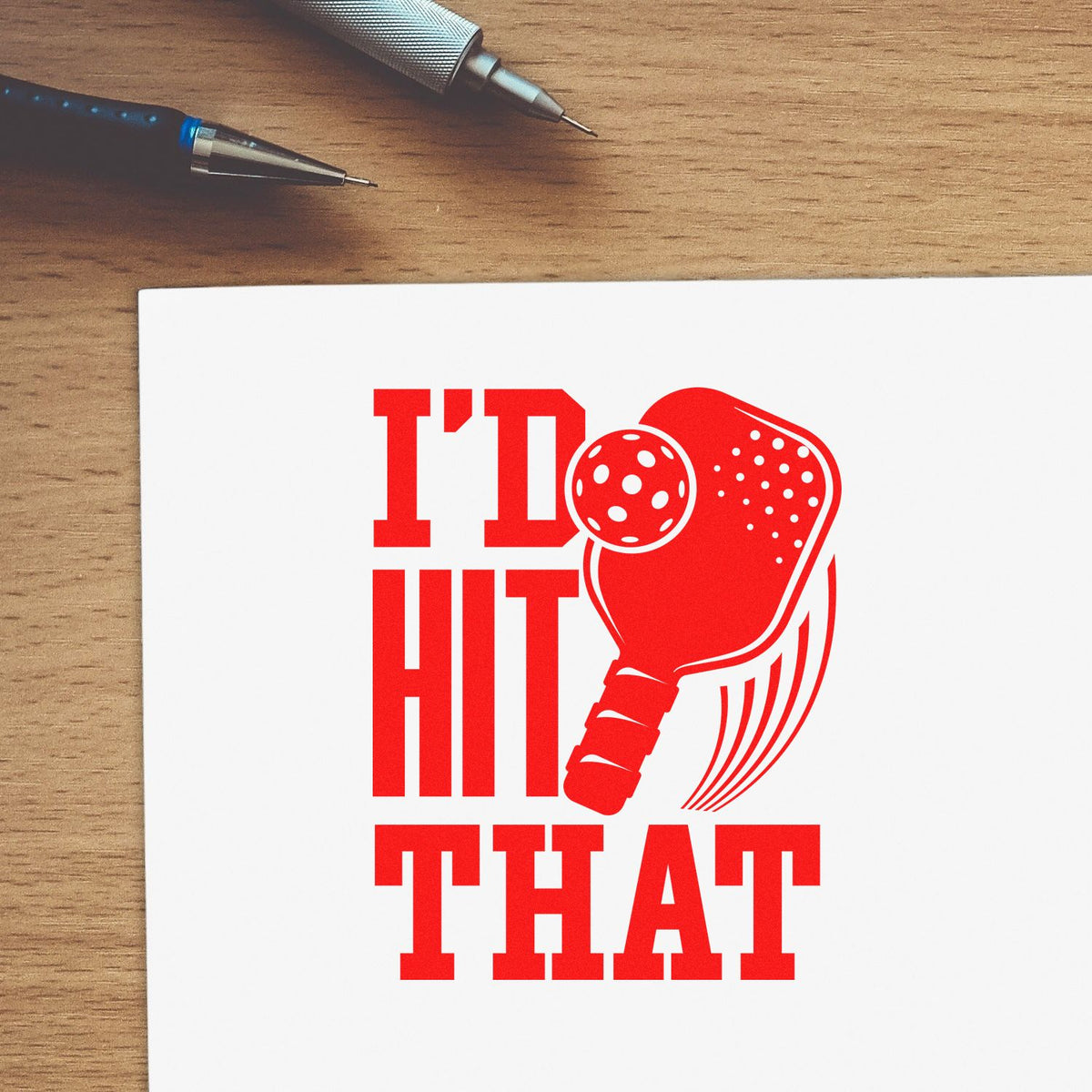 Red pickleball paddle stamp and seal reading Id Hit That on white paper, with pens on a wood desk.