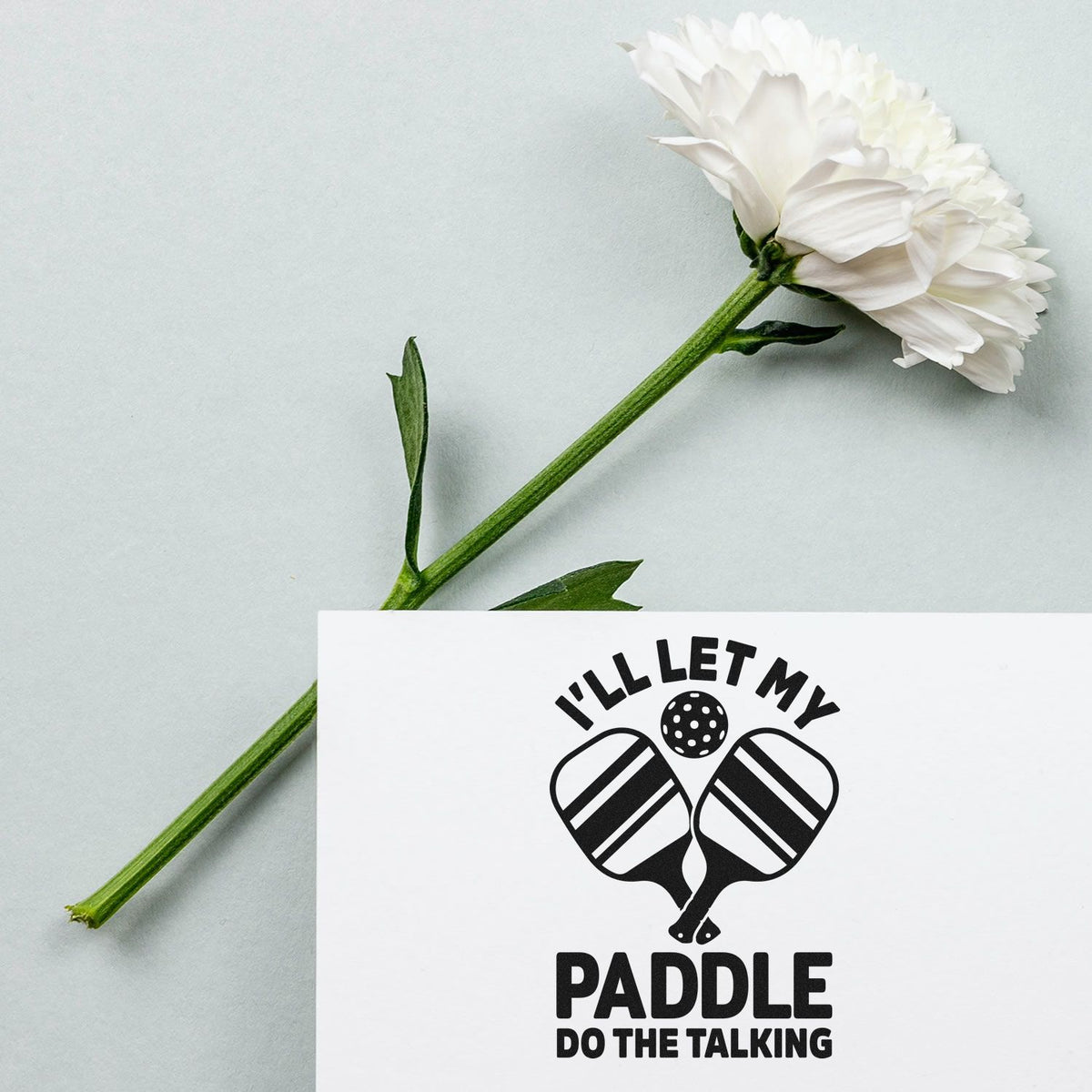 White flower beside a black custom stamp seal reading Ill Let My Paddle Do the Talking with pickleball paddles on a white card.