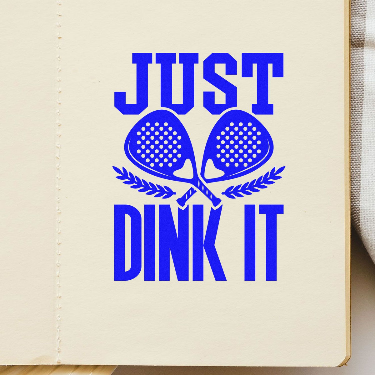Blue stamp seal reading JUST DINK IT with crossed pickleball paddles and laurel on cream paper.