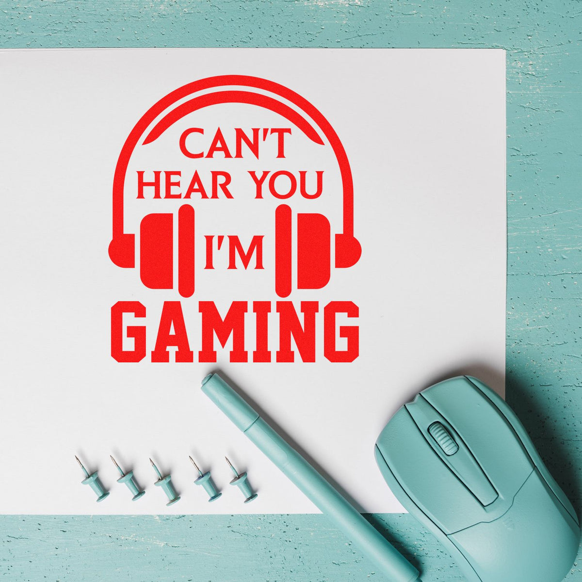 Red rubber stamp design reading Cant hear you Im gaming with headset icon, on white paper beside a teal mouse.